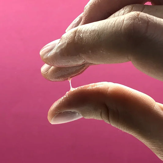 Aufnahme zur Bestimmung der Fruchtbarkeit: Zervixschleim wird zwischen Daumen und Zeigefinger auf seine Spinnbarkeit geprüft. Der Schleim zieht einen deutlichen Faden, was auf die hochfruchtbare Phase im weiblichen Zyklus hindeutet. Pinkfarbener Hintergrund.