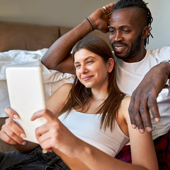 Ein junges Paar liegt entspannt auf einem Bett und schaut gemeinsam auf ein weißes Tablet. Die Szene vermittelt Vertrauen und gemeinsame Entscheidungsfindung beim Thema Familienplanung und natürliche Verhütungsmethoden.