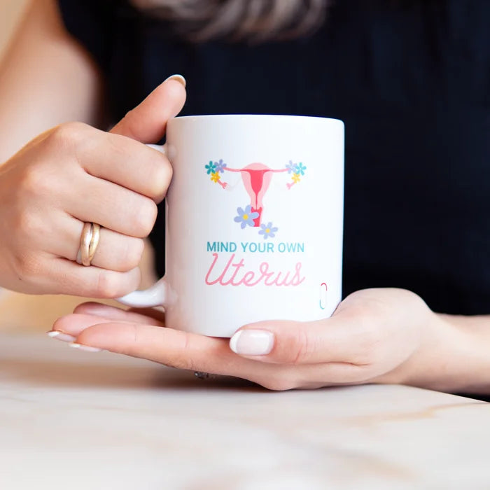 Hände halten eine weiße Kaffeetasse mit der Aufschrift 'MIND YOUR OWN Uterus' und einer blumigen Uterus-Illustration über einem hellen Tisch. Das Design kombiniert blaue und rosa Schriftzüge. Eine authentische Lifestyle-Szene, die das Produkt als modernes Accessoire zeigt.
