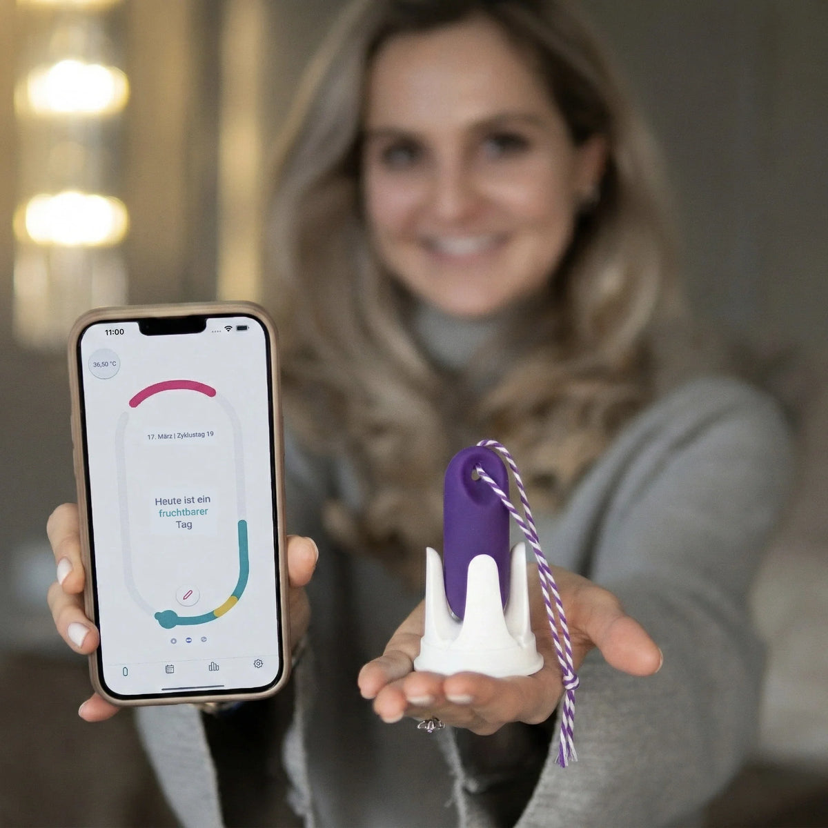 Eine Frau hält lächelnd ein Smartphone mit der geöffneten trackle App und den lila trackleCatch Sensor in die Kamera. Die App zeigt den Status 'Heute ist ein fruchtbarer Tag'. Der Sensor mit Rückholkordel steht in seiner Halterung auf ihrer Handfläche.