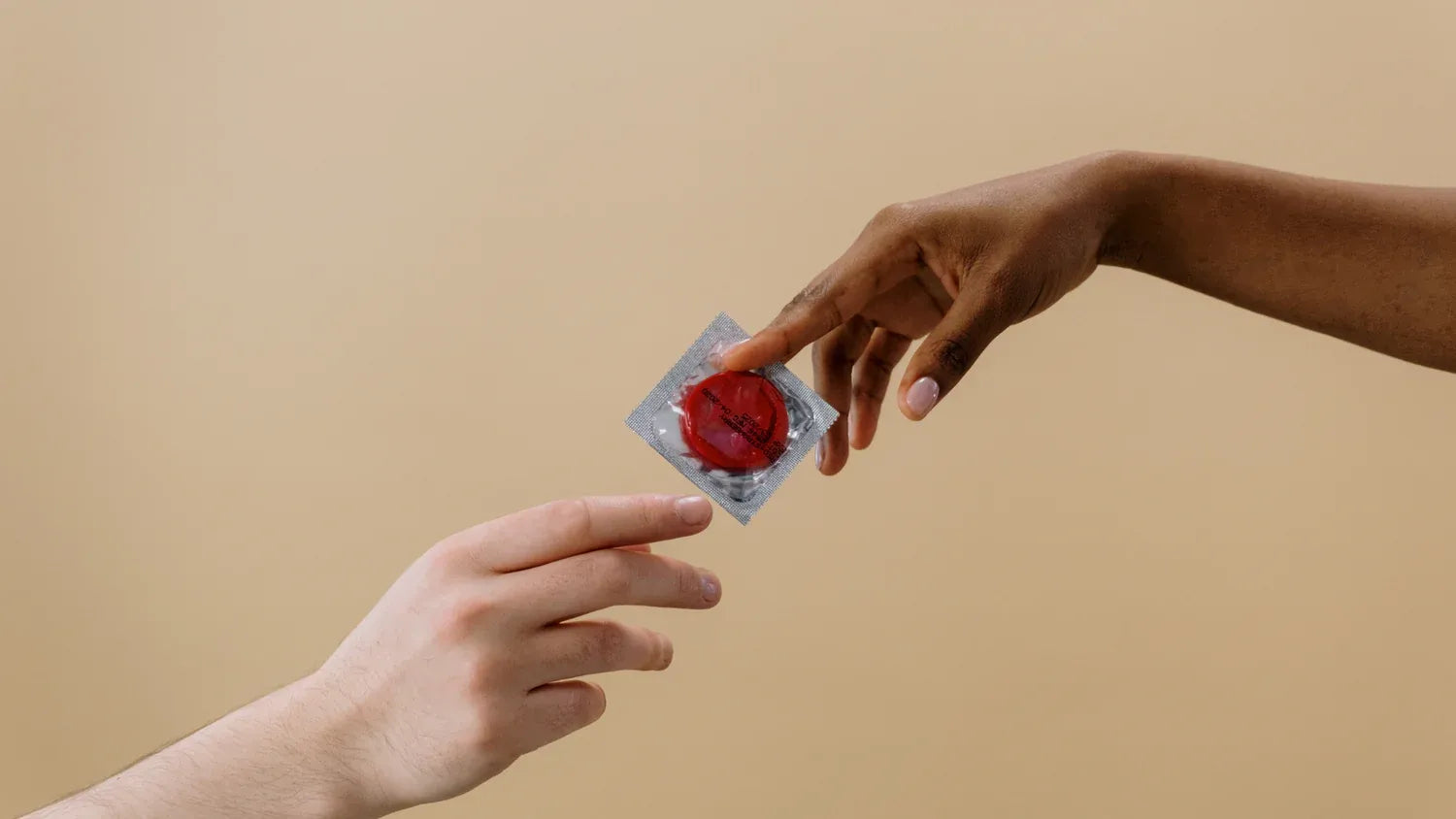 Zwei Hände reichen sich ein verpacktes Kondom vor beigem Hintergrund. Die Geste erinnert an das Motiv 'Die Erschaffung Adams' und symbolisiert Verantwortung und Sicherheit in der Verhütung.
