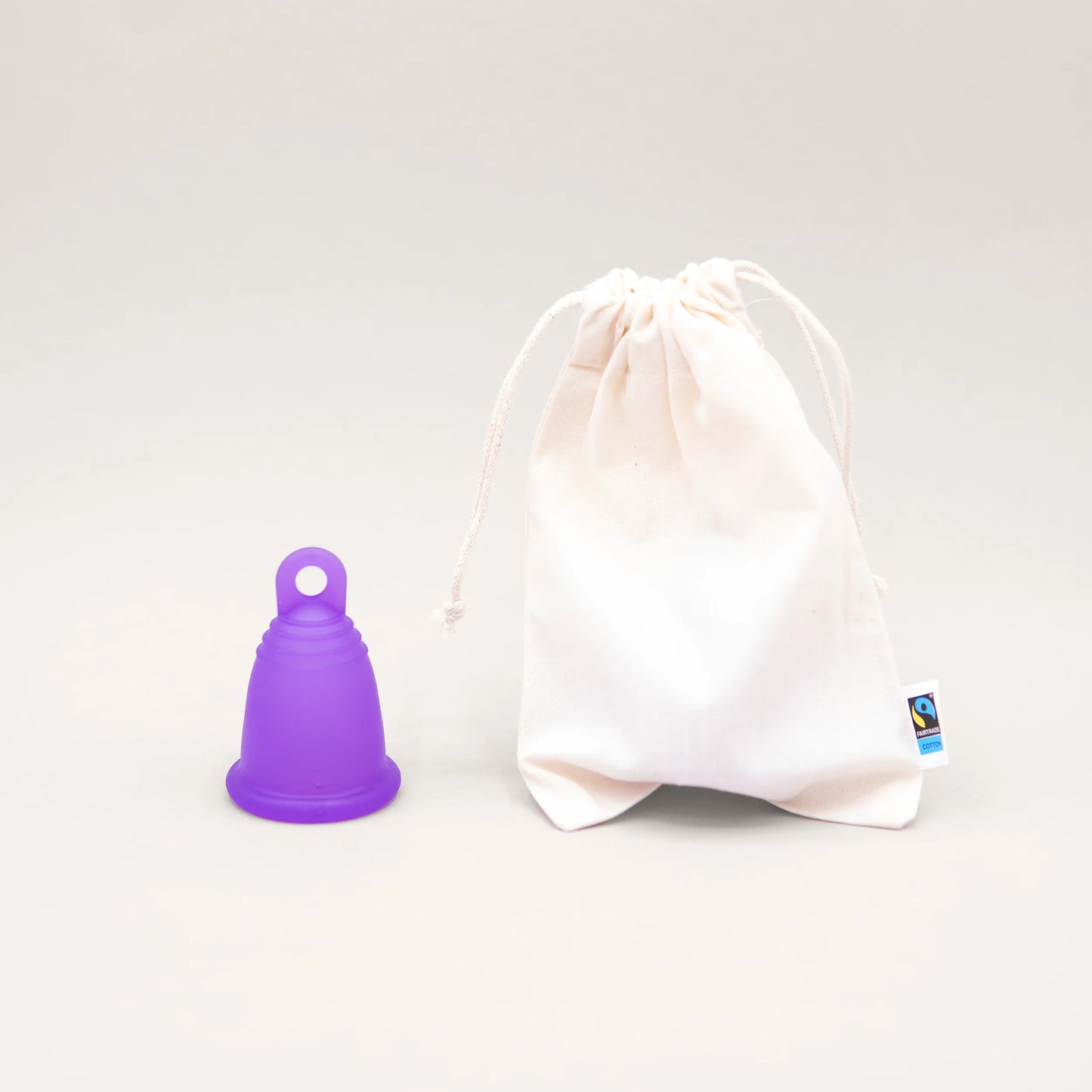 Die lila trackleCup Menstruationstasse steht neben einem weißen Aufbewahrungsbeutel aus Baumwolle mit Kordelzug. Der Beutel trägt das Fairtrade-Cotton-Siegel. Eine nachhaltige und schadstofffreie Alternative zu Tampons und Binden.