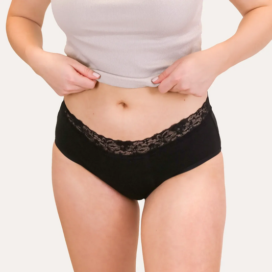Frontale Nahaufnahme der Hipster Strong Periodenunterwäsche in Schwarz. Der elastische Bund ist mit einer breiten, floralen Spitze verziert. Das Model trägt ein hellgraues Ripp-Top, das über den Bund gerafft ist.