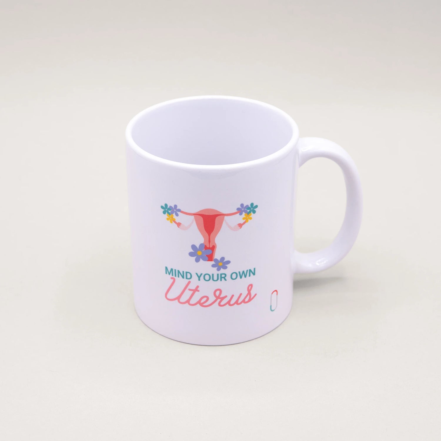 Weiße trackle Tasse, bedruckt mit einer pinkfarbenen Uterus-Illustration, die an den Eierstöcken mit bunten Blumen geschmückt ist. Der Text 'MIND YOUR OWN Uterus' ist in blauer und rosa Schreibschrift darunter platziert.