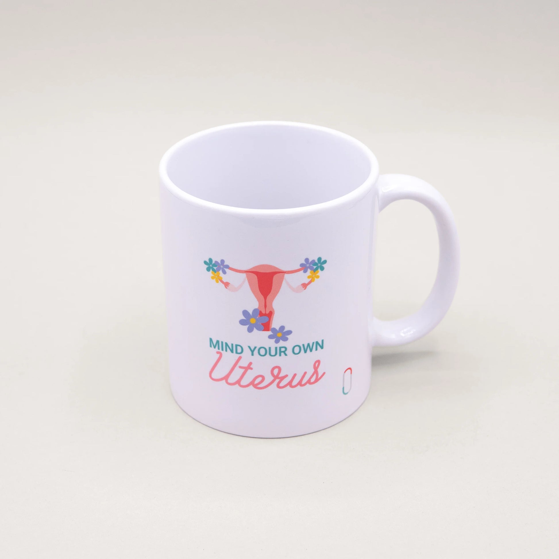 Weiße trackle Tasse, bedruckt mit einer pinkfarbenen Uterus-Illustration, die an den Eierstöcken mit bunten Blumen geschmückt ist. Der Text 'MIND YOUR OWN Uterus' ist in blauer und rosa Schreibschrift darunter platziert.
