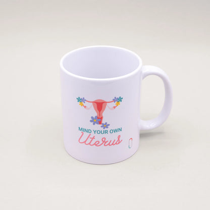 Weiße trackle Tasse, bedruckt mit einer pinkfarbenen Uterus-Illustration, die an den Eierstöcken mit bunten Blumen geschmückt ist. Der Text 'MIND YOUR OWN Uterus' ist in blauer und rosa Schreibschrift darunter platziert.