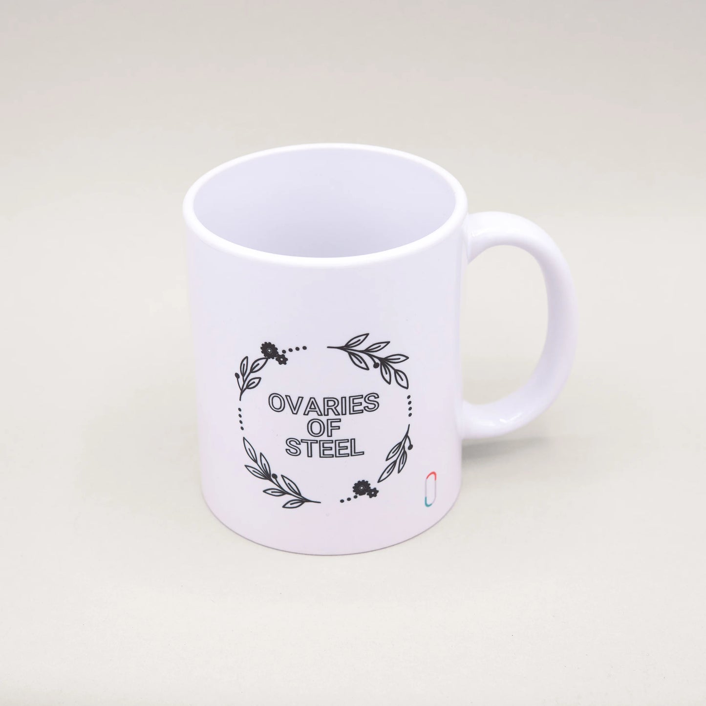 Weiße Tasse mit einem schwarzen Blumen- und Blätterkranz-Design auf der Vorderseite. In der Mitte des Kranzes steht der Spruch 'OVARIES OF STEEL'. Ein dezent plaziertes trackle-Logo ist unten rechts sichtbar.