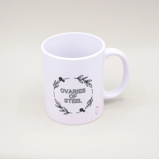 Weiße Tasse mit einem schwarzen Blumen- und Blätterkranz-Design auf der Vorderseite. In der Mitte des Kranzes steht der Spruch 'OVARIES OF STEEL'. Ein dezent plaziertes trackle-Logo ist unten rechts sichtbar.