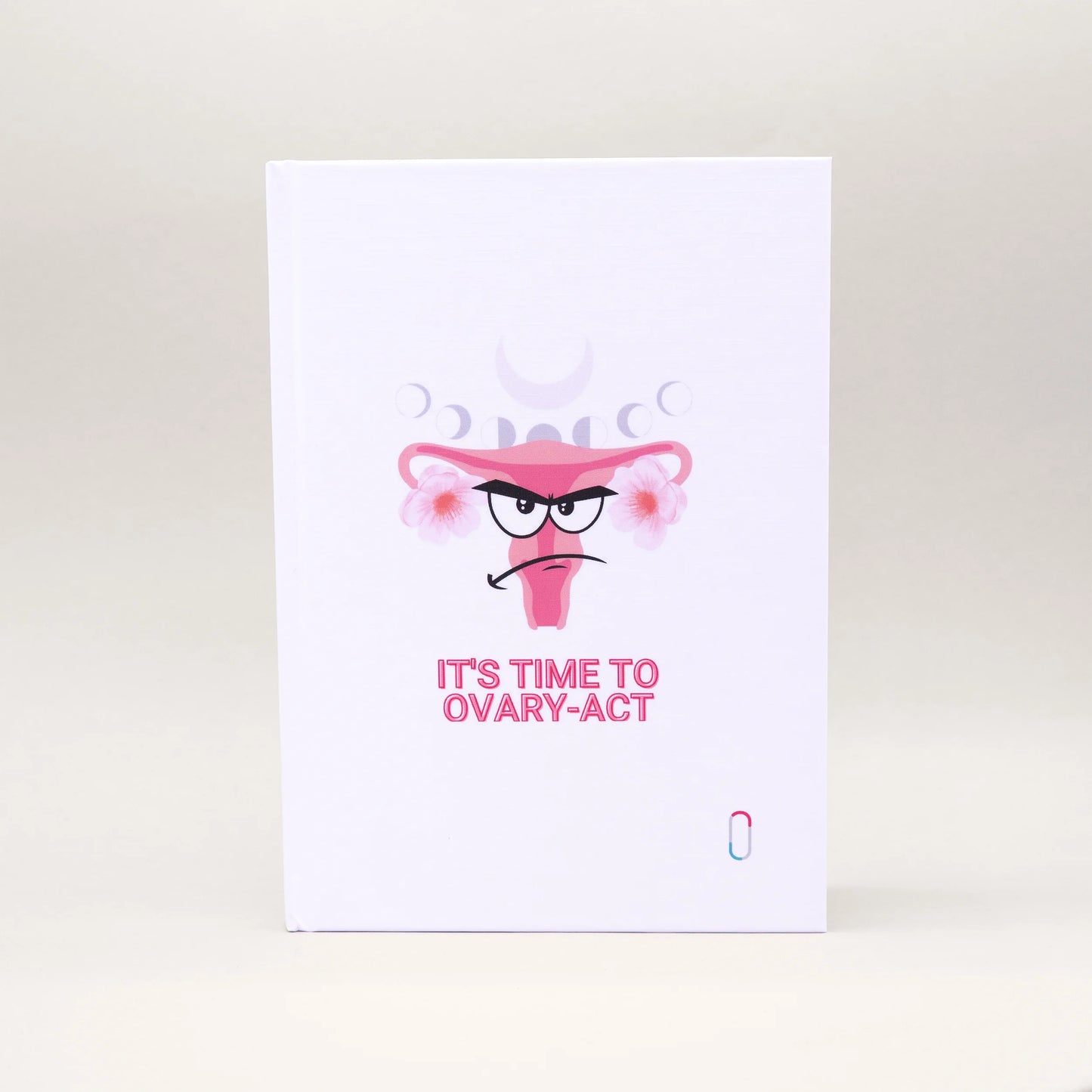Weißes Notizbuch mit einem humorvollen Design: Ein illustrierter Uterus mit grimmigem Gesichtsausdruck (Angry Face) und rosa Blüten. Über der Illustration sind die verschiedenen Mondphasen angedeutet, darunter steht der Spruch 'IT'S TIME TO OVARY-ACT' in rosa Großbuchstaben.