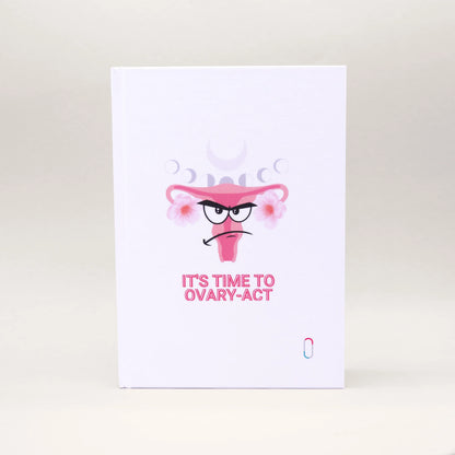 Weißes Notizbuch mit einem humorvollen Design: Ein illustrierter Uterus mit grimmigem Gesichtsausdruck (Angry Face) und rosa Blüten. Über der Illustration sind die verschiedenen Mondphasen angedeutet, darunter steht der Spruch 'IT'S TIME TO OVARY-ACT' in rosa Großbuchstaben.