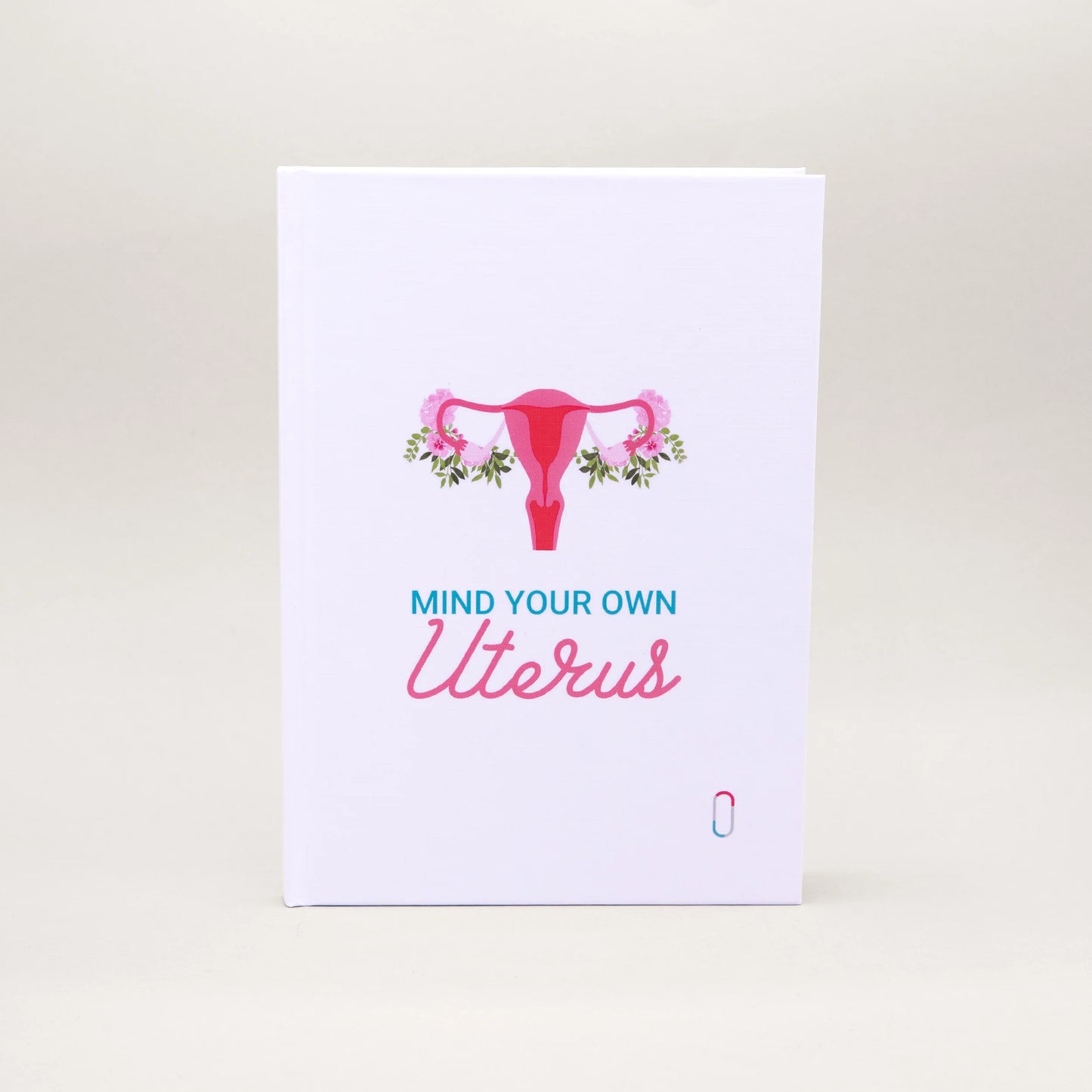 Weißes Notizbuch mit einer pinkfarbenen Illustration eines Uterus, der an den Eierstöcken mit rosa Blumen und grünen Blättern geschmückt ist. Darunter steht in blauer und rosa Schreibschrift der Text 'MIND YOUR OWN Uterus'. Ein modernes Design für Zyklus-Journaling oder Notizen.