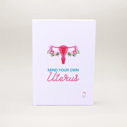 Weißes Notizbuch mit einer pinkfarbenen Illustration eines Uterus, der an den Eierstöcken mit rosa Blumen und grünen Blättern geschmückt ist. Darunter steht in blauer und rosa Schreibschrift der Text 'MIND YOUR OWN Uterus'. Ein modernes Design für Zyklus-Journaling oder Notizen.
