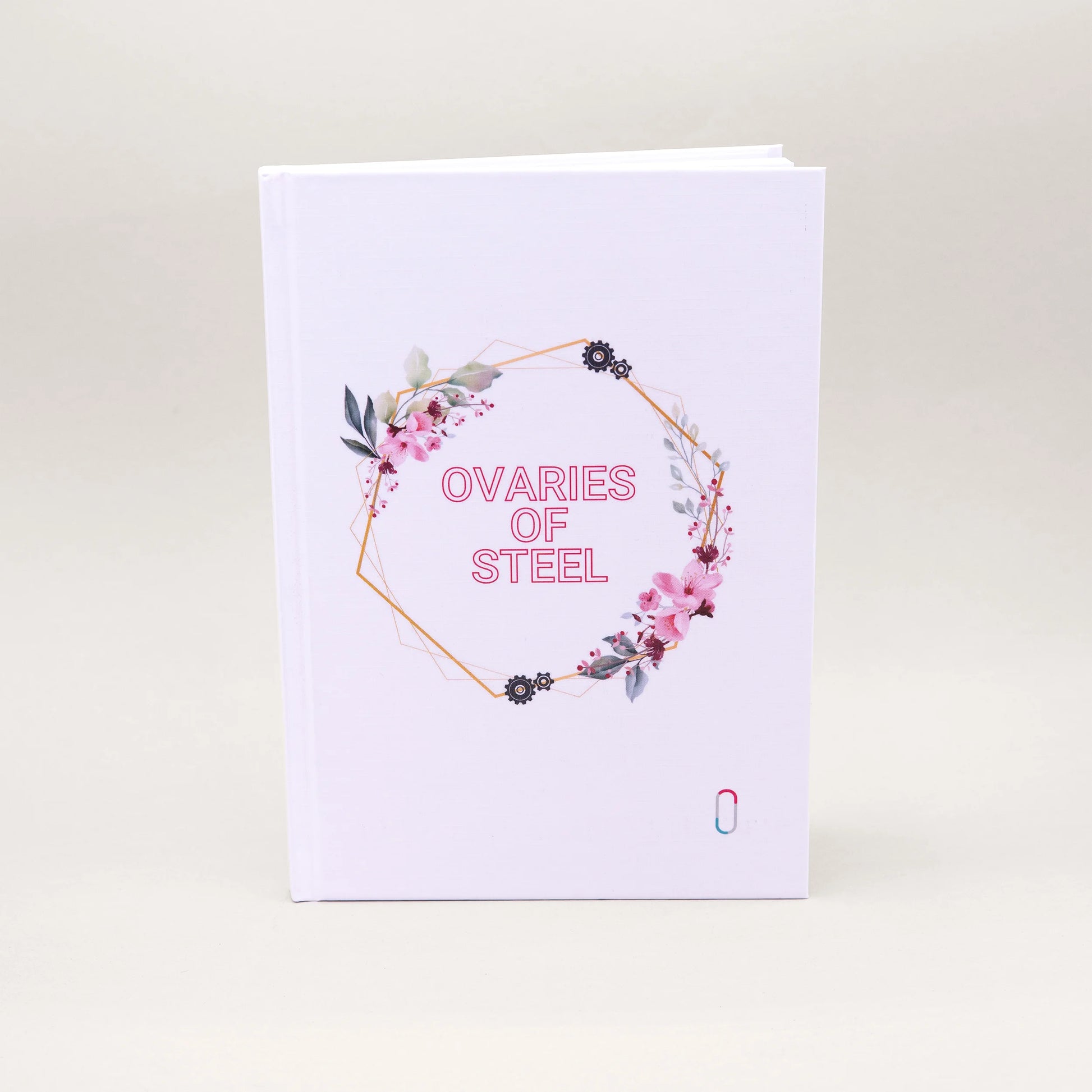 Weißes Hardcover-Notizbuch mit der Aufschrift 'OVARIES OF STEEL' in rosa Buchstaben. Der Text ist von einem geometrischen, goldenen Rahmen umgeben, der mit zarten rosa Blüten, grünen Blättern und kleinen Zahnrad-Illustrationen verziert ist. Unten rechts befindet sich dezent das bunte trackle-Logo.