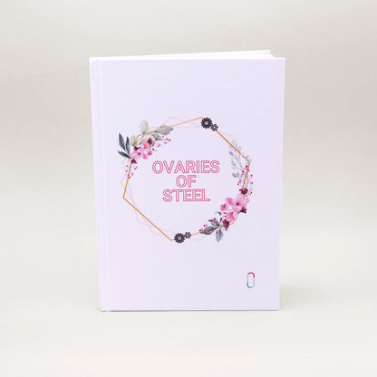 Weißes Hardcover-Notizbuch mit der Aufschrift 'OVARIES OF STEEL' in rosa Buchstaben. Der Text ist von einem geometrischen, goldenen Rahmen umgeben, der mit zarten rosa Blüten, grünen Blättern und kleinen Zahnrad-Illustrationen verziert ist. Unten rechts befindet sich dezent das bunte trackle-Logo.