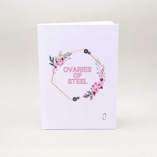Weißes Hardcover-Notizbuch mit der Aufschrift 'OVARIES OF STEEL' in rosa Buchstaben. Der Text ist von einem geometrischen, goldenen Rahmen umgeben, der mit zarten rosa Blüten, grünen Blättern und kleinen Zahnrad-Illustrationen verziert ist. Unten rechts befindet sich dezent das bunte trackle-Logo.
