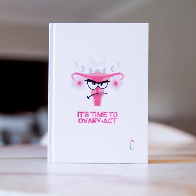 Das weiße Notizbuch 'IT'S TIME TO OVARY-ACT' steht aufrecht auf einer hellen Oberfläche. Der Fokus liegt auf dem Cover-Design mit dem humorvollen Uterus, dem Angry-Face-Gesichtsausdruck, den Mondphasen und dem pinken Schriftzug. Das trackle-Logo ist unten rechts zu sehen. Der Hintergrund ist stark unscharf.