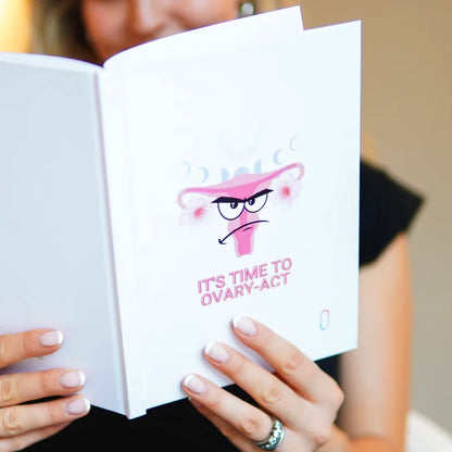 Eine Frau hält das geöffnete Notizbuch 'IT'S TIME TO OVARY-ACT' in beiden Händen und liest darin. Das Cover mit dem Angry-Face-Uterus, den Mondphasen und dem trackle-Logo ist vollständig sichtbar. Der Hintergrund ist unscharf.