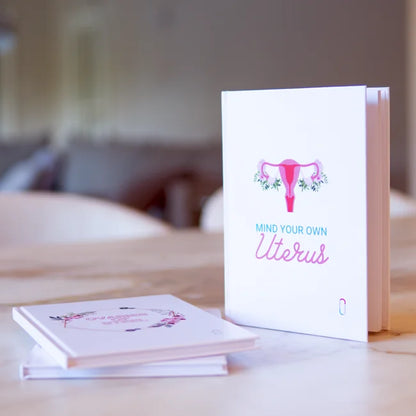 Produkt-Duo am Schreibtisch: Das Notizbuch 'MIND YOUR OWN UTERUS' mit der blumigen Uterus-Illustration steht aufrecht. Im Vordergrund liegen zwei 'OVARIES OF STEEL' Notizbücher flach auf dem Stapel. Die Szene ist hell, mit einem unscharfen Hintergrund.