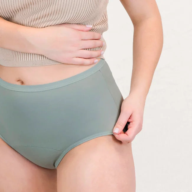 Nahaufnahme der High Waist Periodenunterwäsche in Salbei. Eine Hand zieht leicht am elastischen, flachen Bund an der Hüfte, um die Dehnbarkeit und den weichen Abschluss des Materials zu zeigen.
