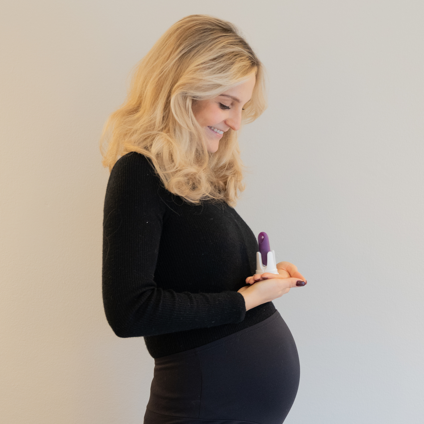 Profilansicht einer lächelnden schwangeren Frau in schwarzer Kleidung, die den lila trackleCatch in seiner weißen Halterung behutsam in den Händen vor ihrem Babybauch hält. Das Bild hat einen hellen Hintergrund und vermittelt eine positive, glückliche Stimmung nach erfolgreicher Familienplanung.