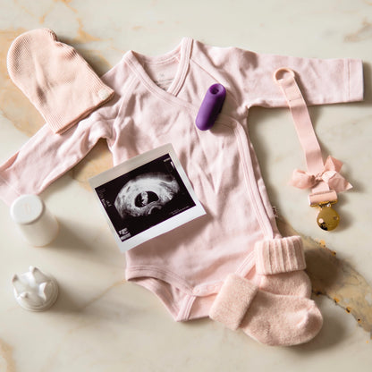 Flachbild-Arrangement (Flatlay) auf Marmorhintergrund: Ein hellrosa Baby-Body mit passender Mütze und Söckchen. In der Mitte liegt ein Schwarz-Weiß-Ultraschallbild einer frühen Schwangerschaft. Direkt daneben ist der lila trackleCatch platziert, links im Bild sieht man die weiße Aufbewahrungsbox und Halterung. Eine elegante Schnullerkette mit goldener Klammer rundet das Bild ab.