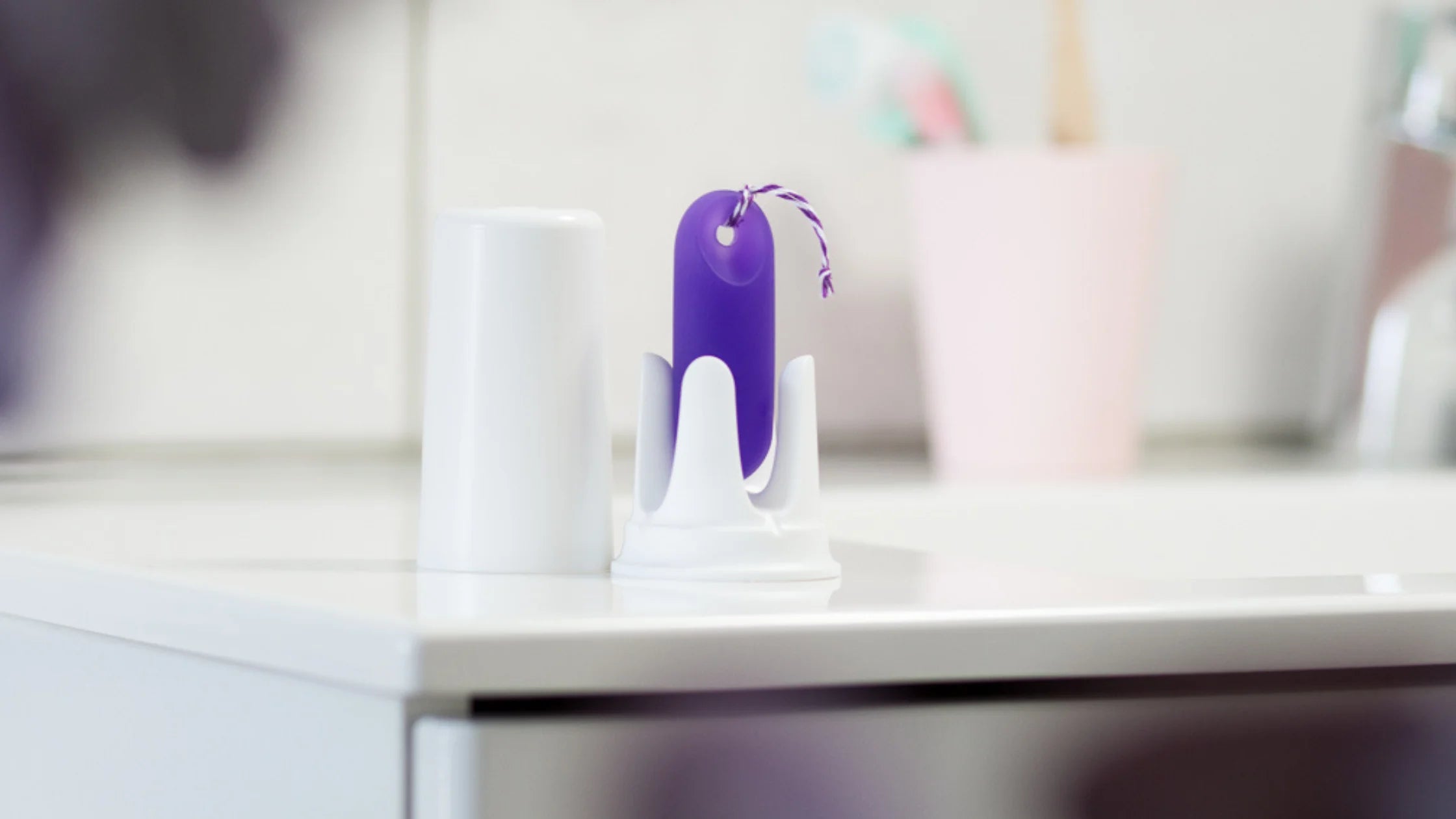 Der lila trackleCatch mit Rückholkordel steht in seiner weißen Halterung auf einem hellen Badezimmerschrank. Im Hintergrund ist unscharf ein rosa Zahnputzbecher zu sehen. Die Szene vermittelt die einfache Integration des Sensors in die morgendliche Routine.