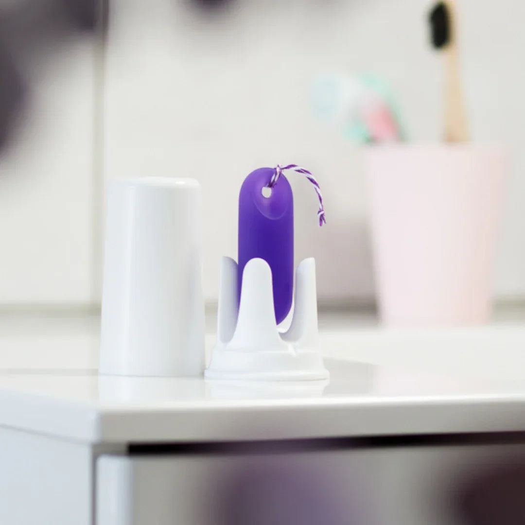 Der lila trackleCatch mit Rückholkordel steht in seiner weißen Halterung auf einem hellen Badezimmerschrank. Im Hintergrund ist unscharf ein rosa Zahnputzbecher zu sehen. Die Szene vermittelt die einfache Integration des Sensors in die morgendliche Routine.