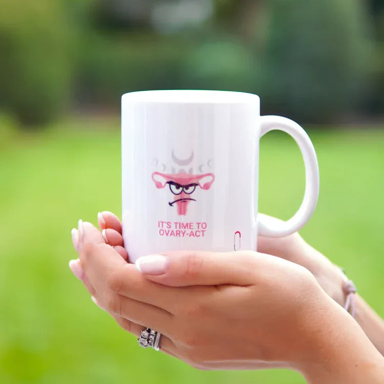 Die trackle Tasse 'IT'S TIME TO OVARY-ACT' wird mit beiden Händen vor einem unscharfen, grünen Gartenhintergrund gehalten. Das humorvolle Uterus-Design mit dem 'Angry Face' und den Mondphasen ist zentral im Bild platziert. Helles, natürliches Tageslicht hebt die Details hervor.