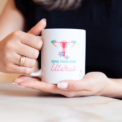 Hände halten eine weiße Kaffeetasse mit der Aufschrift 'MIND YOUR OWN Uterus' und einer blumigen Uterus-Illustration über einem hellen Tisch. Das Design kombiniert blaue und rosa Schriftzüge. Eine authentische Lifestyle-Szene, die das Produkt als modernes Accessoire zeigt.
