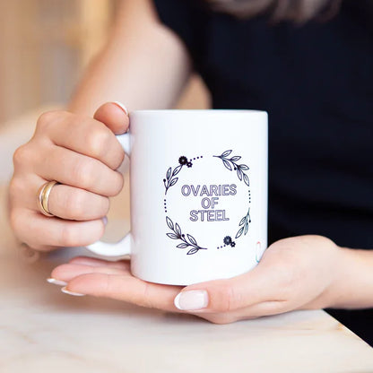 Eine Person hält die weiße trackle Tasse 'OVARIES OF STEEL' mit beiden Händen. Die Hände ruhen auf einer hellen Marmoroberfläche. Das schwarze Blumenkranz-Design der Tasse und das kleine trackle-Logo sind im Fokus.
