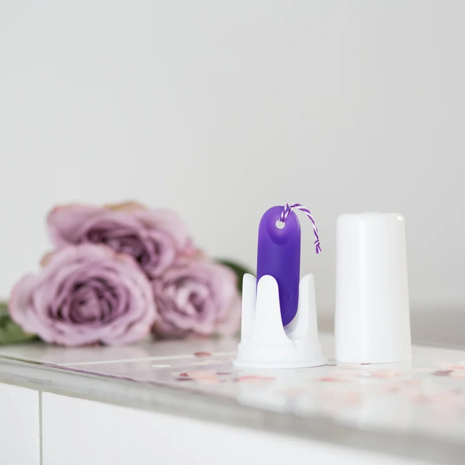 Lifestyle-Foto des trackleCatch Sensors im Badezimmer. Der lila Sensor steht in seiner weißen Aufbewahrungsbox auf einem hellen Fliesenrand, daneben liegen dekorative rosa Rosenblüten. Ein weicher Hintergrund unterstreicht das diskrete Design.