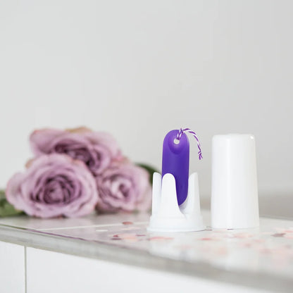 Lifestyle-Foto des trackleCatch Sensors im Badezimmer. Der lila Sensor steht in seiner weißen Aufbewahrungsbox auf einem hellen Fliesenrand, daneben liegen dekorative rosa Rosenblüten. Ein weicher Hintergrund unterstreicht das diskrete Design.