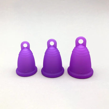Drei lila trackleCup Menstruationstassen in den Größen S, M und L stehen zum Vergleich nebeneinander. Die Cups haben eine ergonomische Form mit einer praktischen Ring-Griffhilfe am Boden für einfaches Entfernen.